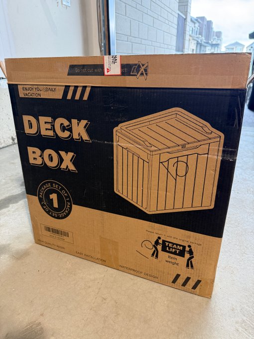 A large cardboard box for a 'DECK BOX' sits on a concrete floor, featuring product details like 'STORAGE SET OF 1', 'WATERPROOF DESIGN', and a 'TEAM LIFT' label.