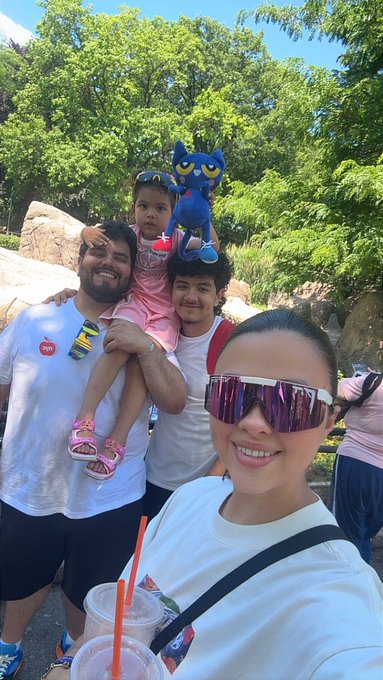 The image shows a group of people posing for a photo, with two men holding their children. The man holding his child has a blue stuffed animal on his shoulder. One person in the group is wearing sunglasses, while another person is carrying an orange straw. The background features trees and rocks, creating a natural setting for this family moment. The caption at the bottom of the image reads "Gm... Day 147 of a selfie a day until MON hits ATH Don't forget to touch grass and enjoy your family and friends!".