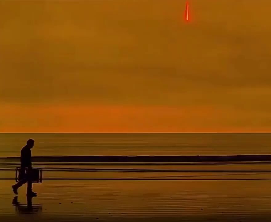 The image shows a man walking along the beach, carrying a suitcase as he walks towards the ocean. There are no other people or objects visible in the scene. The photo has been edited with a red filter applied to it, giving it an artistic and dramatic appearance.