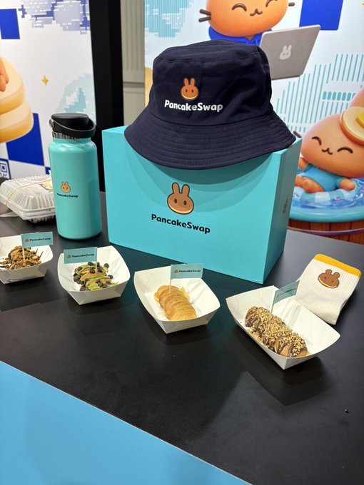A PancakeSwap promotional display featuring a navy bucket hat, a teal water bottle, and various pastries arranged on a table with branded signs.