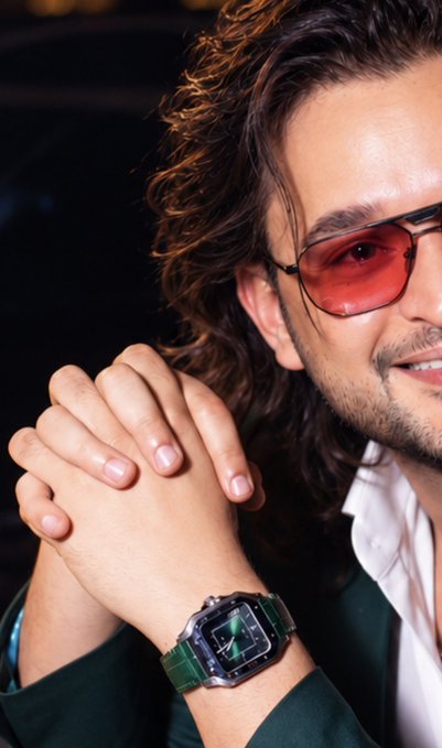A man with curly brown hair and red-tinted aviator sunglasses smiles confidently while clasping his hands together, showcasing a luxury green leather-strapped watch with a tonneau-shaped case. The polished, high-contrast styling and his accessories complement the upbeat "10/10 bull market" mood, radiating the optimistic "bullmorning" energy referenced in the post.