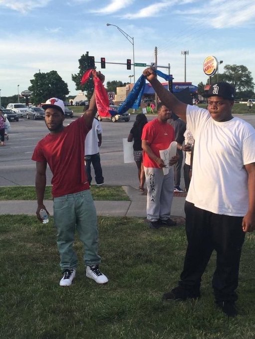 Two young men stand on a grassy area holding up colored bandanas (red and blue), wearing a red shirt with a 'D' cap and a white shirt with a 'D' cap, in what appears to be an urban street setting.