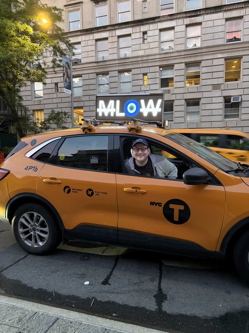 The image features a man sitting inside an orange taxi cab, smiling at the camera. There are two other cars parked nearby, one of which has a sign that reads "Mallow" above its windshield. The scene takes place on a city street with tall buildings in the background and trees lining the sidewalk.