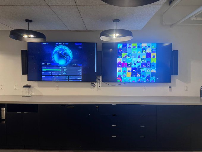 Two flat-screen displays mounted on a wall above dark cabinetry; the left TV shows a video game with a blue circular interface, while the right TV displays a colorful grid of app or video thumbnails.