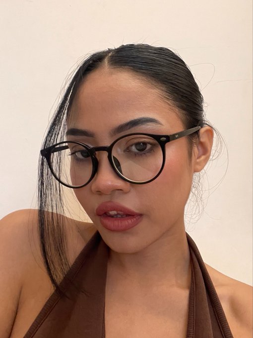A close-up selfie of a young woman with dark hair pulled back, wearing black-framed glasses and a brown halter top.