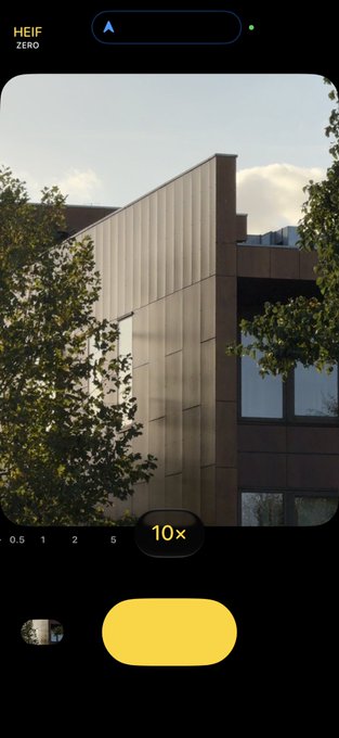 A smartphone camera app interface labeled "HEIF ZERO" displays a 10x zoom view of a modern building with a paneled facade partially framed by green trees, with the post noting the shot was taken at 10x magnification in the new Zerocam app.
