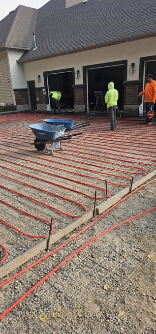 Workers install a hydronic snow melt system in a residential driveway, laying out 3600 sq ft of red 5/8" pex tubing spaced at 12 inches on rebar over compacted gravel, with a wheelbarrow and materials staged nearby. The installation, which will be filled with a water/glycol mix and connected to a Bitcoin mining-heated heat exchanger, demonstrates the on-site preparation before concrete is poured over the tubing grid.