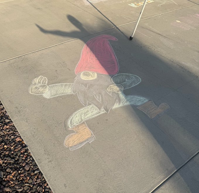A chalk drawing of a garden gnome with a red pointed hat, outstretched arms, and boots on a concrete surface, partially covered by shadows.