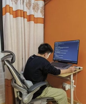 A person wearing a black shirt sits at a desk in a room with orange walls, working on code displayed on a monitor while using an ergonomic chair.