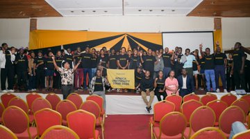 The image shows a large group of people gathered together for an event at a venue with red chairs arranged around a stage. The crowd consists of students from Africa University, posing for a photo. A banner can be seen hanging above them, possibly displaying information about the event or the university itself. The atmosphere appears to be celebratory and positive as everyone seems to be enjoying themselves.