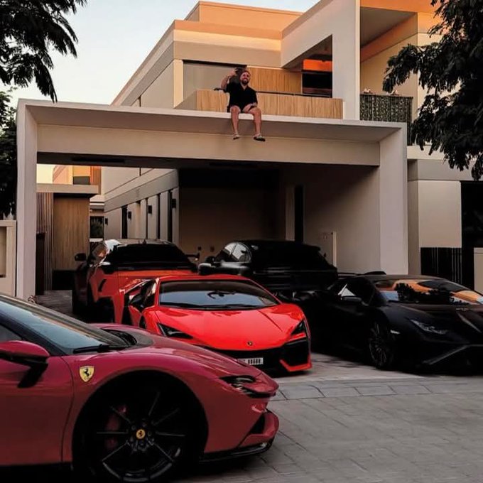 The image features a man sitting on top of his red sports car, which has a black stripe down the middle. The car is parked next to other cars, including a black sports car and a red sports car. A person can be seen standing in front of the white house with a balcony that overlooks the parking lot where these cars are parked.