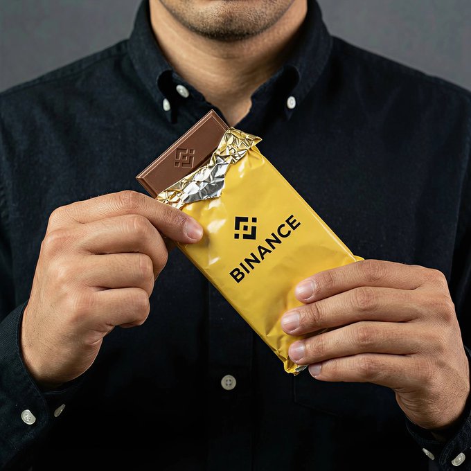 A man in a dark button-up shirt holds a gold-wrapped chocolate bar with a bite taken out, revealing the chocolate stamped with the Binance logo and the brand name "BINANCE" printed on the wrapper—tying into the post celebrating #WorldChocolateDay with Binance-branded chocolate.
