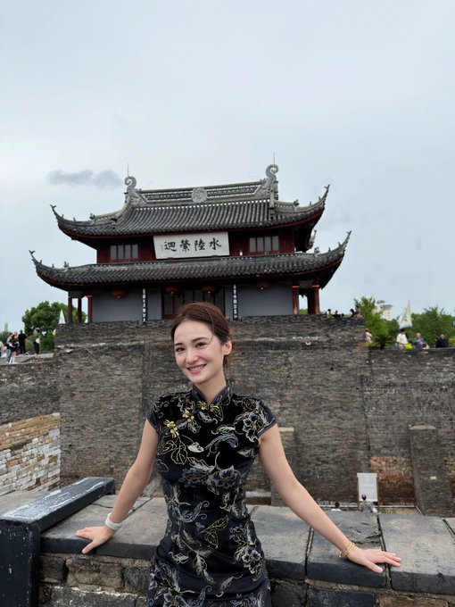 A young woman wearing a floral cheongsam (qipao) dress posing and smiling in front of a traditional Chinese gate tower featuring '水陆演迴' (Shui Lu Yan Hui) calligraphy.