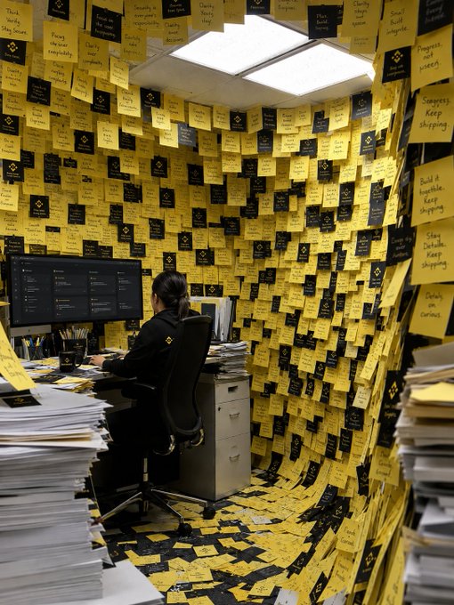 A person works at a computer in a tiny office completely covered floor-to-ceiling in yellow and black sticky notes bearing reminders like "Stay Focused," "Keep Shopping," and "Build a Stable," embodying the "do not disturb" concentration level through an overwhelming visual barrage of tasks.