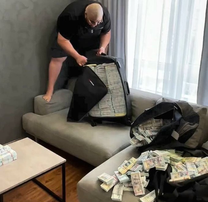 A bald man in a black shirt and shorts stands on a gray couch, unzipping a large suitcase stuffed full of bundles of U.S. dollar bills, with additional stacks of cash spilling out of another bag and scattered across the cushions.