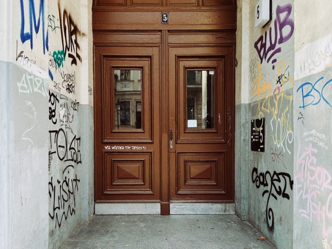 A pair of brown wooden double doors with the number 3, set within a doorway alcove heavily covered in graffiti tags on both side walls.