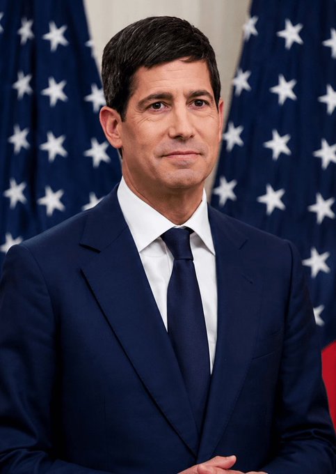 A portrait of a man in a dark suit and tie standing in front of American flags, appearing to be an official government photo.