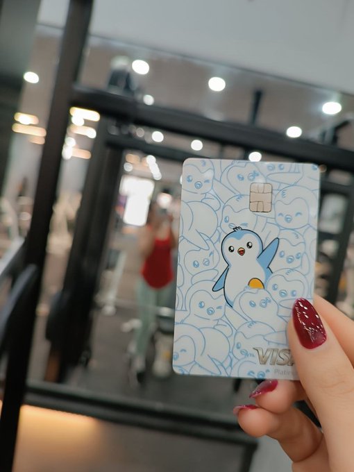 A hand with dark red nail polish holds up a Visa credit card decorated with a blue cartoon penguin design, with a blurred indoor scene in the background.