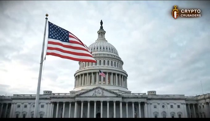 The U.S. Capitol building with the American flag flying in the foreground under a cloudy sky, with a 'CRYPTO CRUSADERS' logo in the upper right corner.