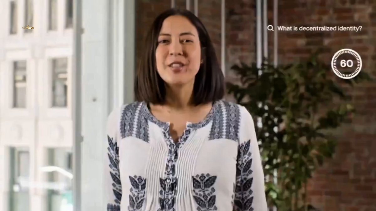 The image features a woman wearing a white blouse with black floral patterns, standing against a brick wall. The background shows a window that allows natural light into the room. There are also some cars visible outside of the building through the window.