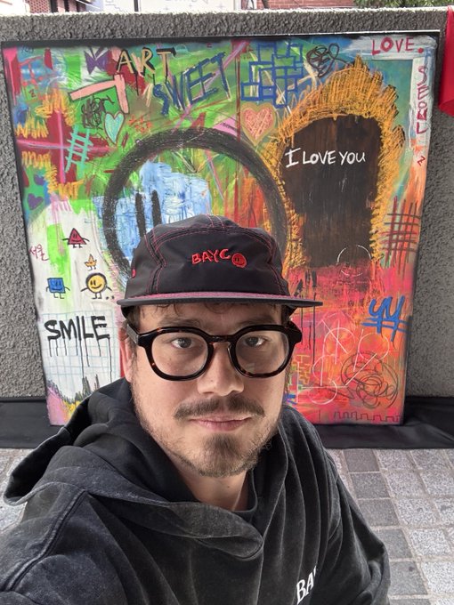 A man wearing black-rimmed glasses and a dark cap with red 'BAYC' embroidery poses in front of a colorful graffiti artwork featuring the words 'ART IS SWEET', 'SMILE', and 'I LOVE YOU'.