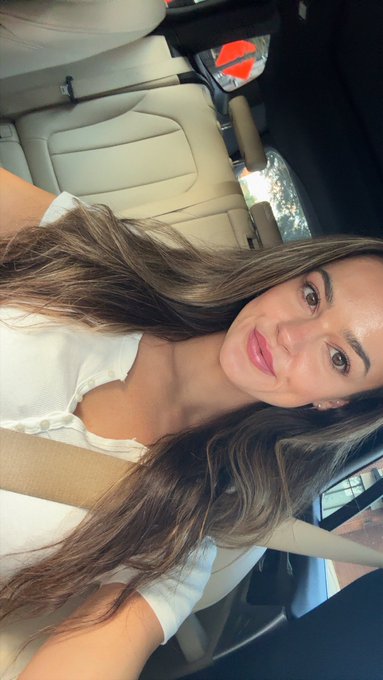 A woman with long brown hair wearing a white top sits in a car, smiling at the camera.