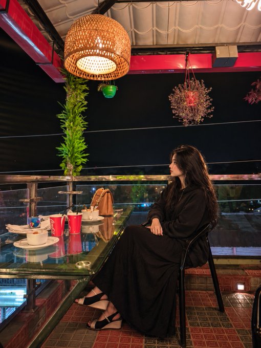 A woman with long dark hair, wearing a black abaya and black sandals, sits at a glass table in a dimly lit restaurant with woven pendant lights and decorative plants.