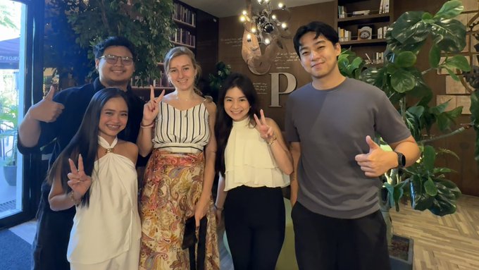 The image shows a group of five people posing together for a photo, with one person wearing a white dress and another wearing a black shirt. The group appears to be enjoying their time at an event or gathering, as they are all smiling and giving thumbs up. The background features a potted plant and some bookshelves, suggesting that the location might be a library or a similar indoor setting. The caption for this image reads: "Dash is in Manila Sat down with amazing people today. They know this market inside out and were generous enough to share it all with me. I came to learn. I stayed for the friendship."