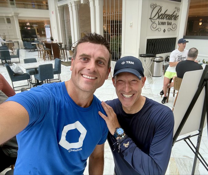 Two men pose for a selfie in what appears to be a hotel lobby or lounge area, with one wearing a blue Chainlink t-shirt and the other wearing a dark long-sleeve shirt and a black TRM cap; a 'Botero Lobby Bar' sign is visible in the background.