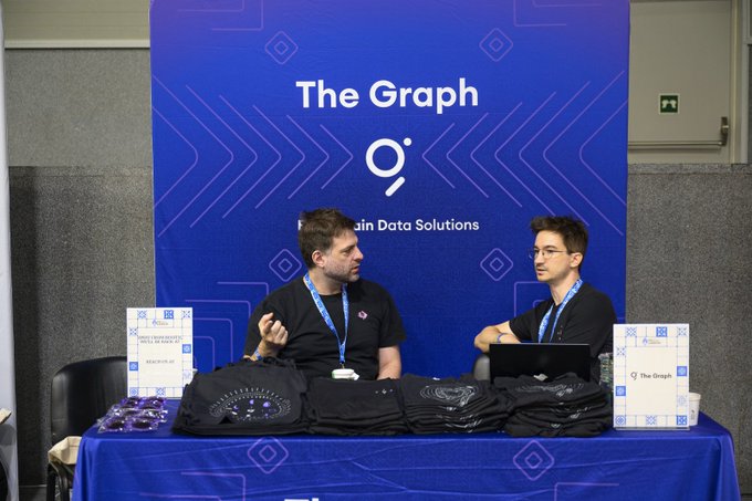 Two men sit at a "The Graph" booth promoting their blockchain data solutions at ETHGlobal Lisbon, where $15,000 in prizes is being offered for the best AI tooling, AI use case, and standardized Graph products. The booth features branded merchandise, promotional materials, and laptops as representatives engage with attendees.