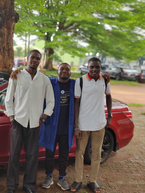 Three young men pose together with their arms around each other in front of a red car, with trees and parked vehicles in the background.