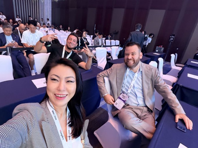 A group of conference attendees smile for a selfie, with one man in the background photobombing by making peace signs. The lanyards read "Yong," "Arthur," and the smiling man in the gray suit holds a badge labeled "ALEX." The post humorously calls out the playful photobomber who crashed the shot.