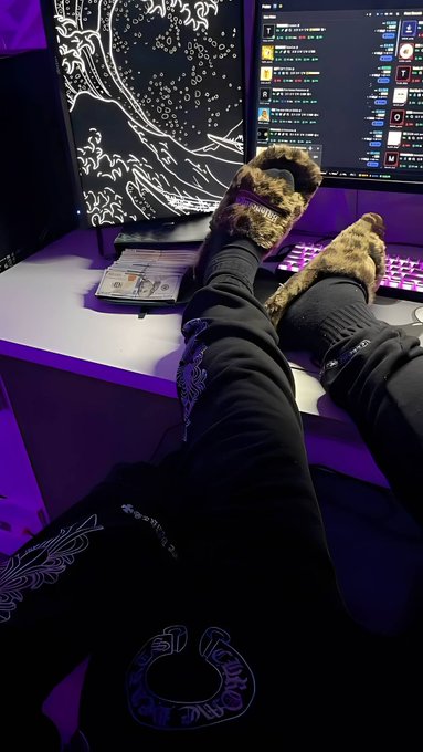 A first-person perspective photo of feet wearing fuzzy leopard-print slippers propped up on a white desk in front of two computer monitors displaying artwork and a trading interface, with an open notebook nearby.