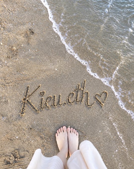 A person stands barefoot on a sandy beach, with the name "Kieu.eth" written in the sand beside a small heart, just as gentle waves wash ashore.