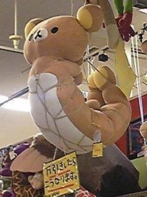 A slightly damaged, light brown Rilakkuma bear plushie hanging in a store display, accompanied by a small yellow Japanese warning sign.