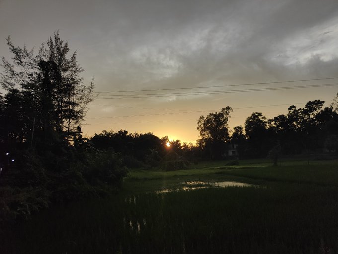A serene landscape photograph captures a sunset over a rural scene with silhouetted trees, a small pond, and green fields under a cloudy sky, with power lines crossing the horizon.
