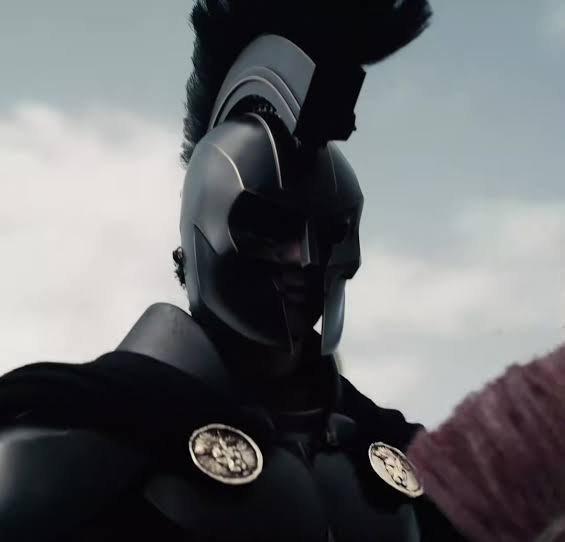A close-up of a warrior wearing a dark Greek-style helmet with a black plume, clad in black armor with circular medallions, set against a cloudy sky.