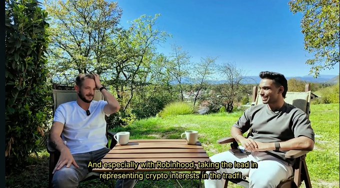 Two men seated outdoors at a wooden table in a sunny setting, with subtitles reading 'And especially with Robinhood, taking the lead in representing crypto interests in the trade.'