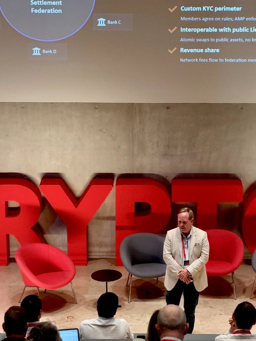 The image shows a man standing behind a podium with a large screen displaying information about cryptocurrency custody services. There are several people sitting in chairs around him, possibly attending a conference or event related to cryptocurrency. The room has a gray wall featuring red letters spelling out "RYPT".