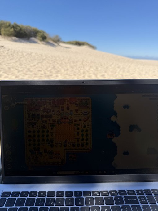 A laptop displaying a pixelated 2D video game map sits on a sandy beach with grassy dunes and a clear blue sky in the background.