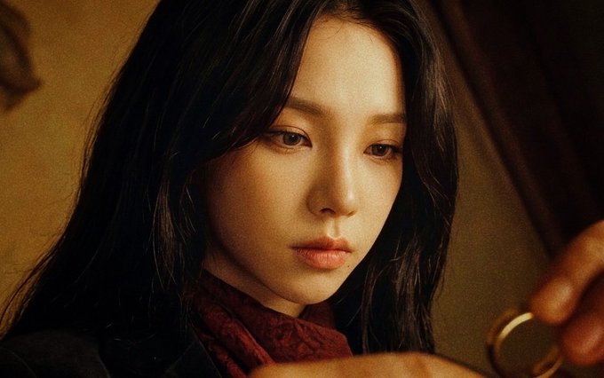 A cinematic close-up of a young woman with long dark hair, wearing a dark top and red scarf, gazing intently at something in her hand under warm lighting.