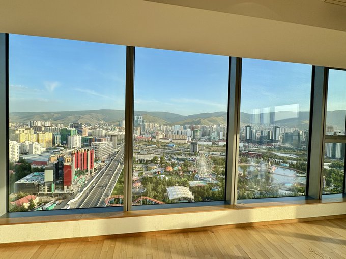A cityscape is viewed through large windows, showing buildings, a road, and mountains under a clear blue sky.