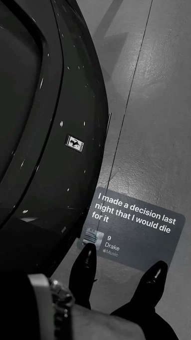 A black-and-white selfie showing black boots on a tiled floor next to a dark bag, with an overlaid Spotify-style music caption reading 'I made a decision last night that I would die for it' by Drake.