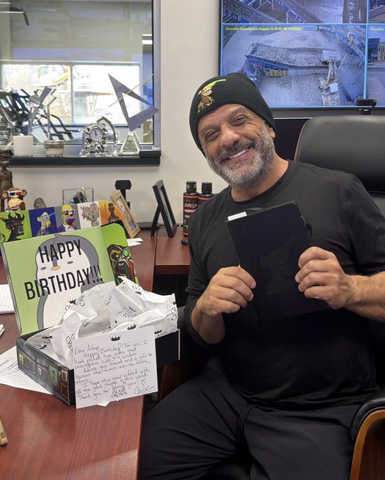 A man wearing a Bored Ape beanie smiles while holding a black journal, with a "HAPPY BIRTHDAY!!!" card and handwritten note from @ImGlassCrown on his desk.