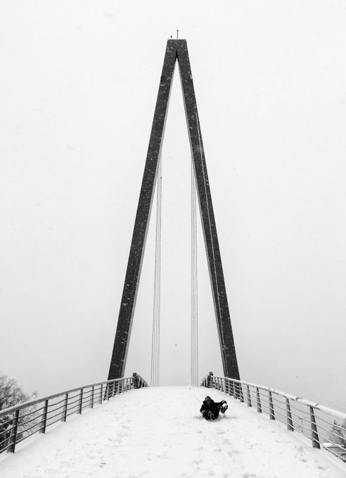 A lone figure sits on a snow-covered pedestrian bridge that leads toward a towering A-frame pylon, surrounded by fog and softness. The image evokes the post's theme of "I believe in my art," capturing the solitary, introspective journey of a creator facing an enormous, almost otherworldly structure.