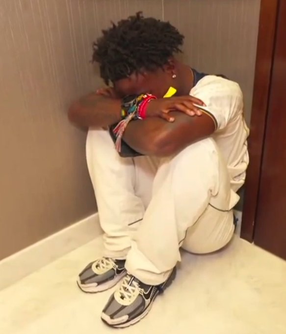 A young man with curly hair sits on the floor in the corner of a room, head resting on his knees in a contemplative pose, wearing white sweatpants, a white t-shirt, and gray Nike sneakers. The image evokes a somber or reflective mood that contrasts with the casual "Gm CT" (Good morning Connecticut or Crypto Twitter) greeting, suggesting a humorous juxtaposition of a "tough morning" or emotional moment against the upbeat salutation.