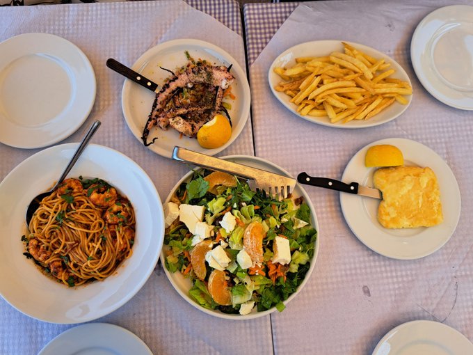 A restaurant table is set with multiple dishes including spaghetti with shrimp, grilled fish with lemon, French fries, a Greek salad, and breaded cutlets, with empty plates ready for diners.
