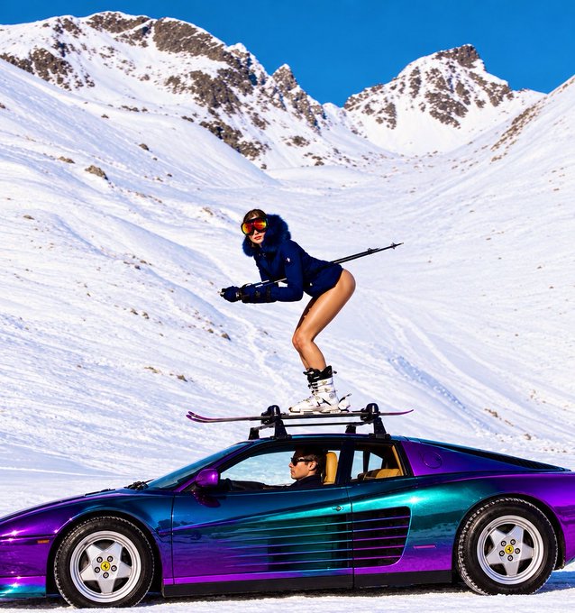 A woman in a blue ski jacket stands on skis strapped to the roof rack of a purple and teal sports car parked in a snowy mountain landscape, with a driver visible inside.