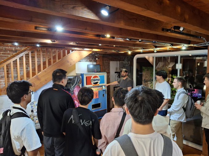 A group of young people gathered indoors around a blue PEPSI vending machine, with one person pointing at it.