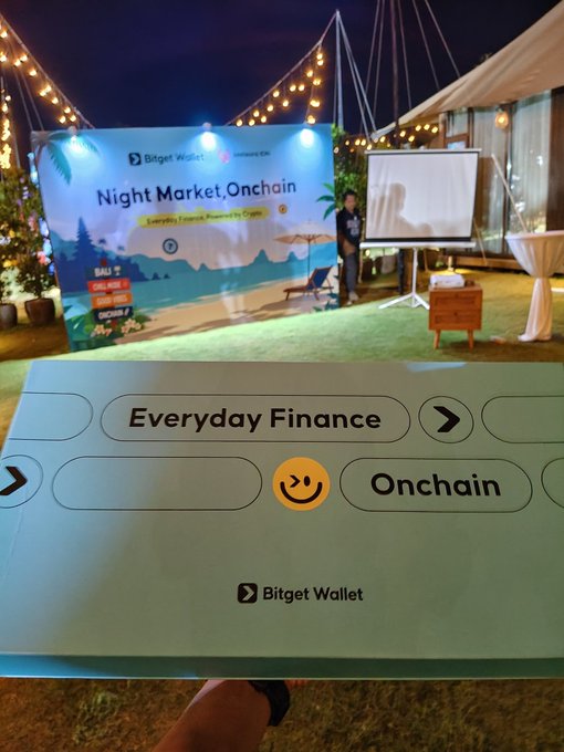 A hand holds a 'Bitget Wallet' branded card at a 'Night Market, Onchain' event by 'Everyday Finance Reunion by CGVN,' with a 'BAL> 5' box visible in the background.